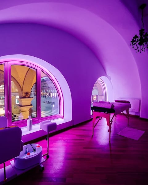 Modern futuristic interior with vibrant purple neon lighting, arched ceiling, glowing window, massage table, and minimalist furniture